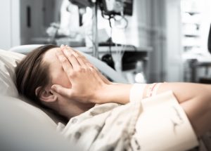 Young woman patient lying in hospital bed with hands over her face, seems distressed